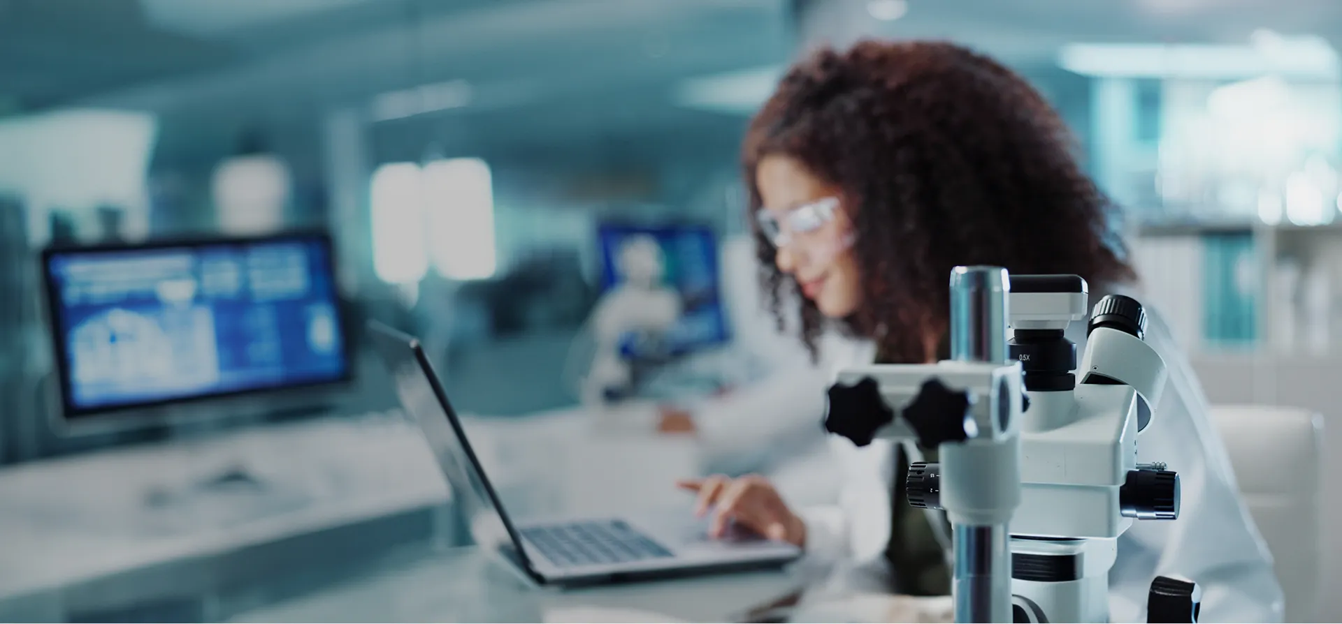 先端技術が並ぶ明るいラボにて、顕微鏡越しにノートパソコンでデータを確認する女性研究員の姿。