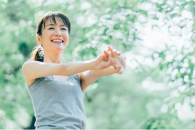 柔らかな木漏れ日の中で、笑顔で腕を伸ばしストレッチをする女性。健康、活力、前向きな姿勢をイメージさせる屋外の風景。