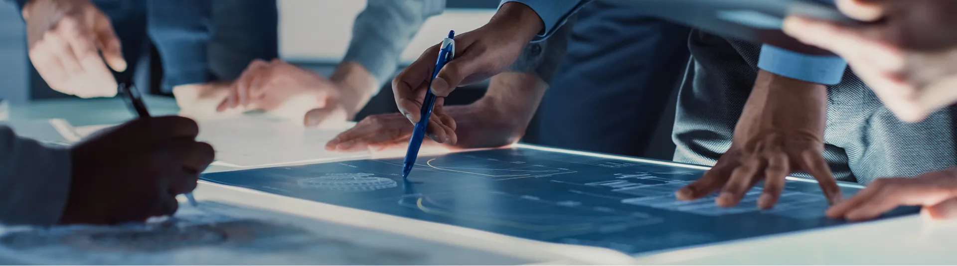 青く光るデジタル設計図を囲み、複数のメンバーがペンを手に議論や検討を行っている様子。
