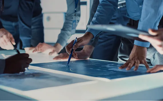 青く光るデジタル設計図を囲み、複数のメンバーがペンを手に議論や検討を行っている様子。